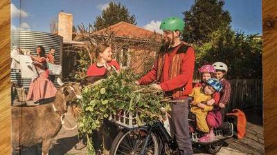 cover of the book Retrosuburbia by David Holmgren featuring a smiling man on a bike with three kids on the back, and a basketfull of greens in the front bike basket; he's smiling at a woman holding a baby goat, and another goat is standing in front of the bike nibbling the greens. A house, water tank, a man, and a woman wearing a baby on her back are in the background.