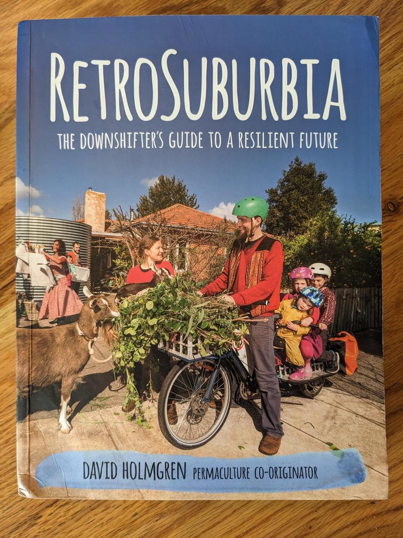 cover of the book Retrosuburbia by David Holmgren featuring a smiling man on a bike with three kids on the back, and a basketfull of greens in the front bike basket; he's smiling at a woman holding a baby goat, and another goat is standing in front of the bike nibbling the greens. A house, water tank, a man, and a woman wearing a baby on her back are in the background.