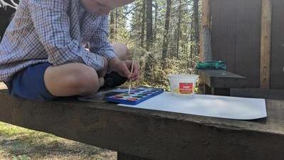 a boy in a button-up shirt sits cross-legged on a wooden bench outdoors,leaning over a tray of watercolors and a blank piece of paper, holding a paintbrush