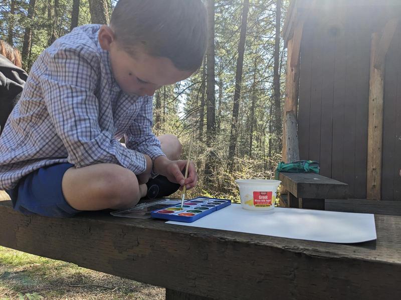 a boy in a button-up shirt sits cross-legged on a wooden bench outdoors,leaning over a tray of watercolors and a blank piece of paper, holding a paintbrush