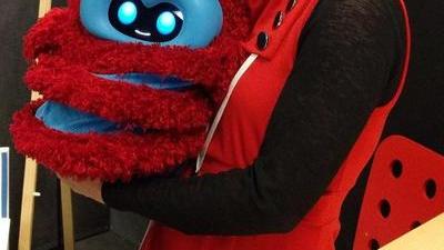 Jacqueline wearing a red dress smiling and holding a robot the size of a large pumpkin; the robot is fuzzy and red with blue stripes and blue plastic around its black and blue face