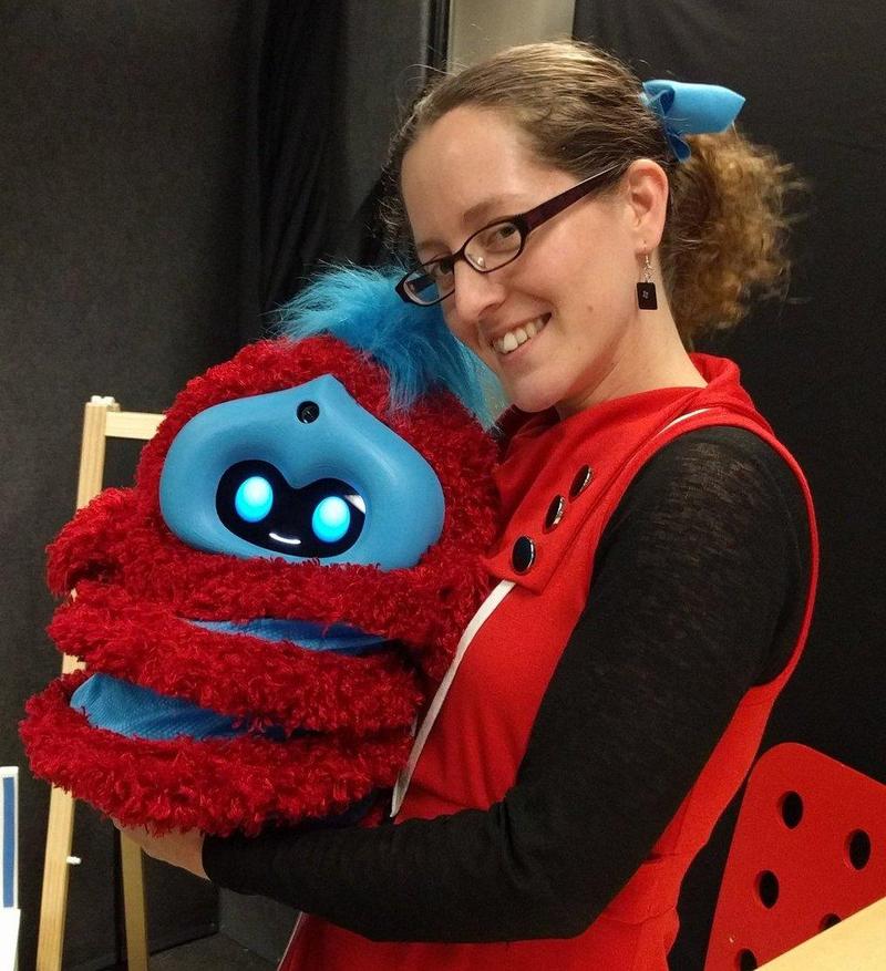 Jacqueline wearing a red dress smiling and holding a robot the size of a large pumpkin; the robot is fuzzy and red with blue stripes and blue plastic around its black and blue face