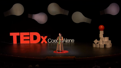 Jacqueline standing on a red carpet circle on stage in front of big letters saying TEDx Couer d'Alene