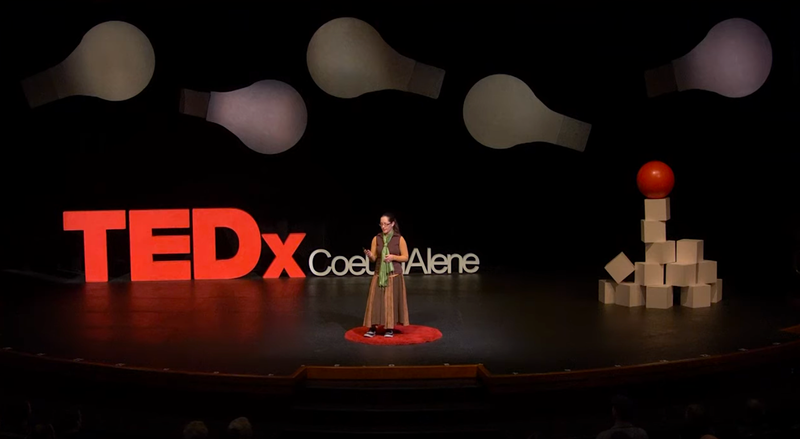 Jacqueline standing on a red carpet circle on stage in front of big letters saying TEDx Couer d'Alene