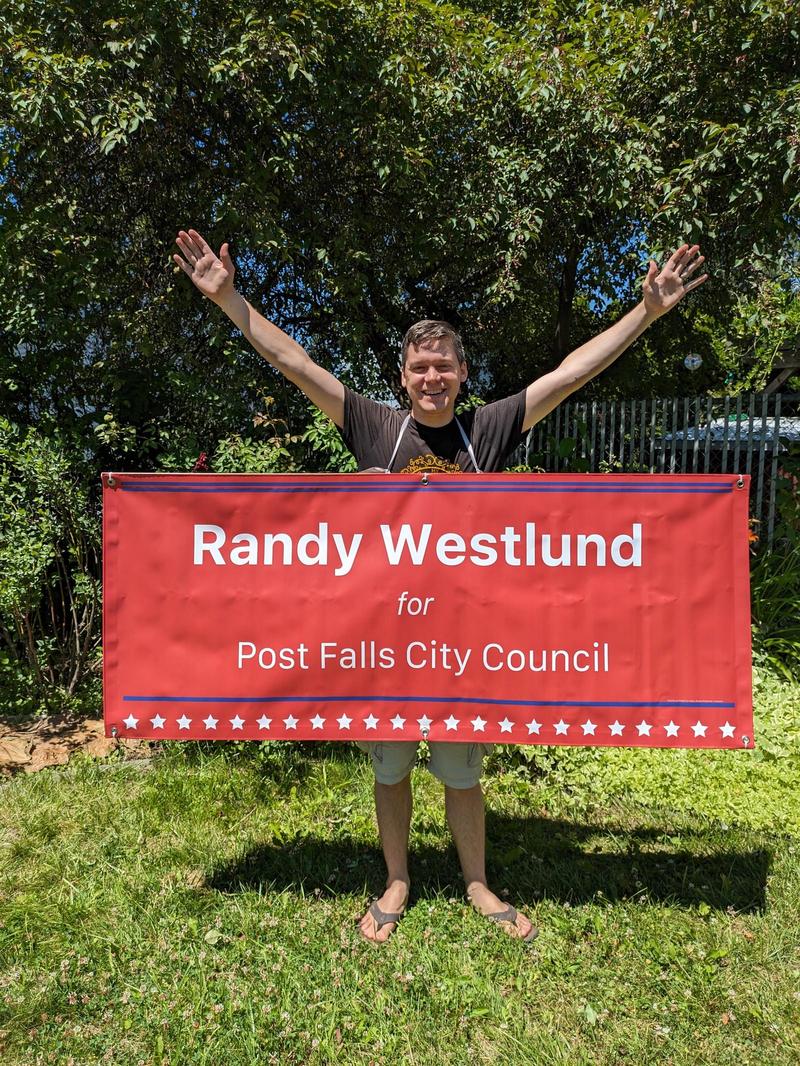 Randy standing in a lawn holding a large banner that says Randy Westlund for Post Falls City Council