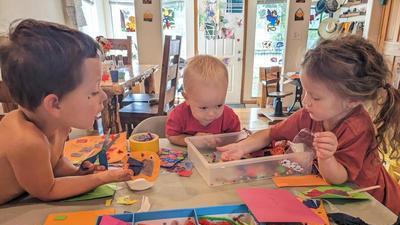 three kids sitting on three sides of a square table leaning in, doing crafts: the table is covered in brightly colored paper, foam, scissors, goggly eyes, and so forth.