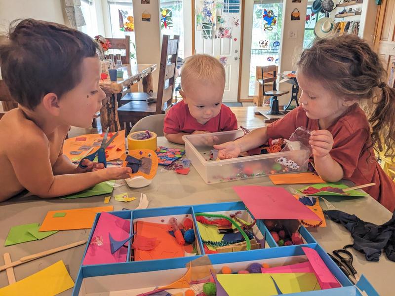 three kids sitting on three sides of a square table leaning in, doing crafts: the table is covered in brightly colored paper, foam, scissors, goggly eyes, and so forth.