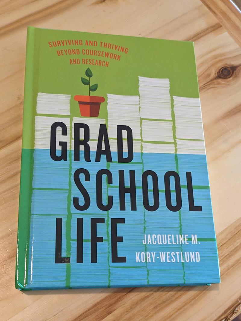 hardcover book Grad School Life: Surviving and Thriving Beyond Coursework and Research by Jacqueline M. Kory-Westlund, resting face up on a table. The cover shows a piles of papers behind the title, with a small potted plant on top of one stack, and the bottom half of the page covered in blue as if underwater