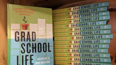 The book Grad School Life: Surviving and Thriving Beyond Coursework and Research by Jacqueline M. Kory-Westlund, standing up on a bookshelf with a stack of more copies behind. The cover shows a piles of papers behind the title, with a small potted plant on top of one stack, and the bottom half of the page covered in blue as if underwater