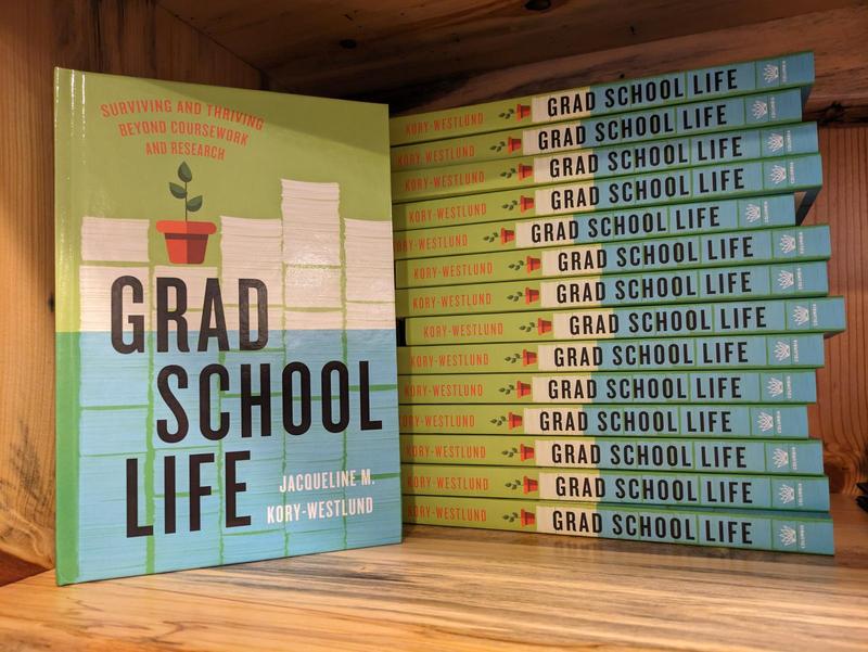 The book Grad School Life: Surviving and Thriving Beyond Coursework and Research by Jacqueline M. Kory-Westlund, standing up on a bookshelf with a stack of more copies behind. The cover shows a piles of papers behind the title, with a small potted plant on top of one stack, and the bottom half of the page covered in blue as if underwater
